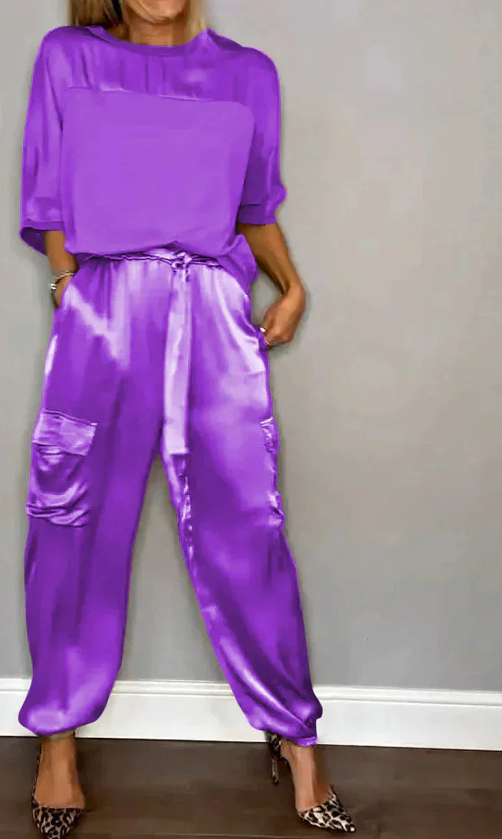 Damen Satin Jumpsuit in Lila, ausgestattet mit Taschen und Bindegürtel, aus weichem, glänzendem Satinmaterial gefertigt.